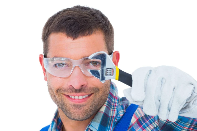 Plumber wearing safty glasses holding an adjustable spanner like a magnifying glass to his eye